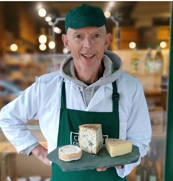 Profile picture of Manager Chris holding a selection of cheese outside the shop in uniform; white lab coat, green apron and cloth hat, with white Chorlton cheesemongers logo