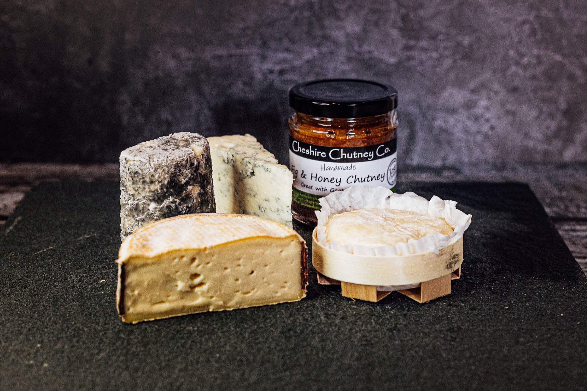 4 delicious looking artisanal cheeses on a slate, showing a mix of colours, shapes and textures, with an additional jar of honey